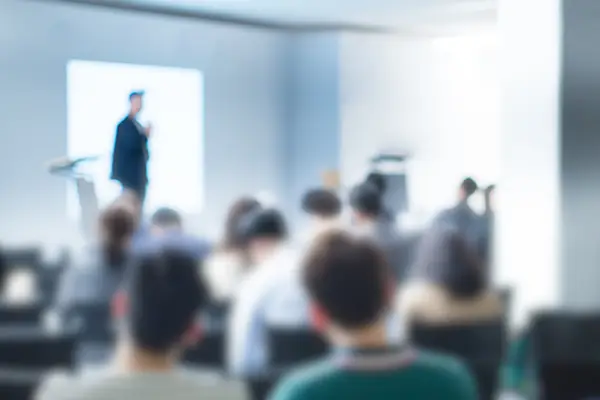 Blurred/defocused speaker at end of room in front of audience in presentation room